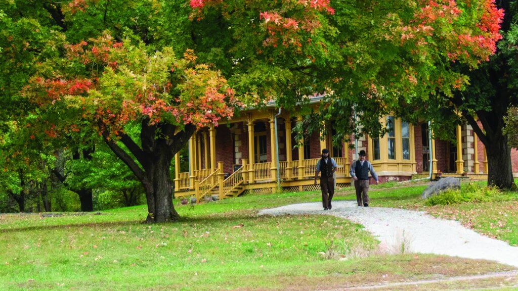 two men in historic clothing walking in front of flynn mansion under fall-colored trees